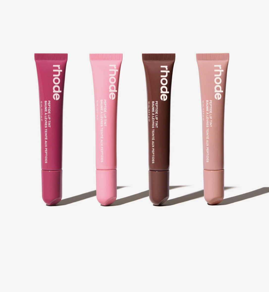 Four Rhode lip balm tubes in different shades of pink, mauve, brown, and nude, arranged in a row on a light gray background with soft shadows.