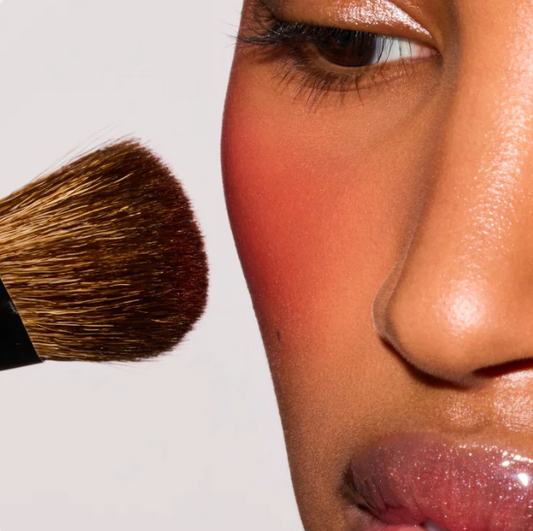 Close-up of a person's face with warm pink and peach makeup, a makeup brush at the left edge, and a clean light gray background.