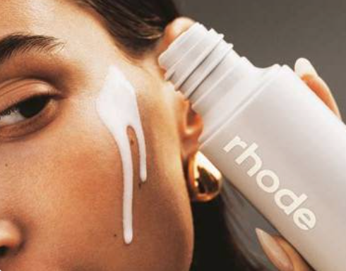 A close-up of a person applying a white cream from a white Rhode cosmetic bottle onto their cheek.