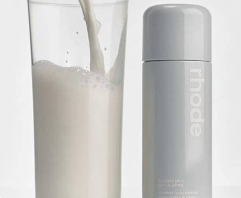 Rhode Glaciers Milk Face Cleansing bottle in gray shown next to a glass of milk being poured, on a light gray background.