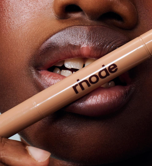 A close-up of a person holding a Rhode liquid makeup stick across their lips. The product is a warm beige/nude shade, and the brand name 'rhode' is clearly printed on the barrel in black.