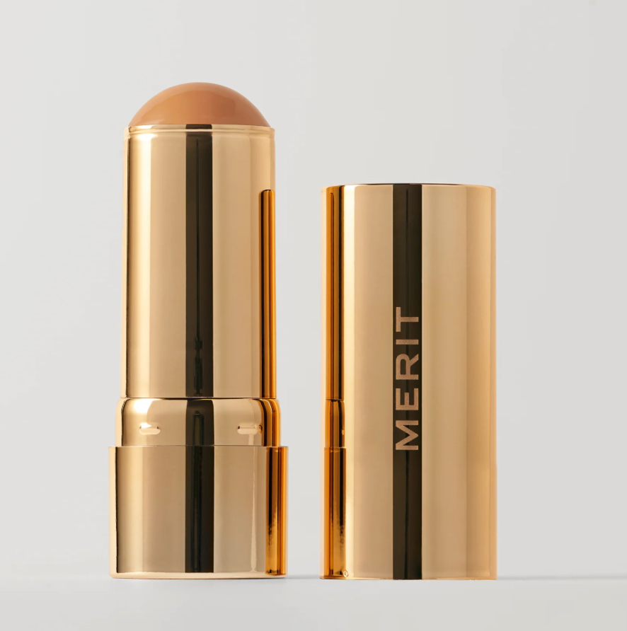 A gold cosmetic stick product shown open next to its matching cap on a light gray background. The exposed product tip is a warm tan shade, and the cap features the brand name MERIT in gold lettering.