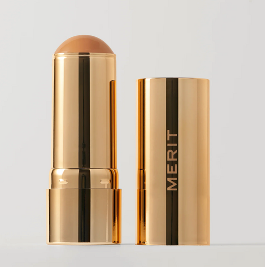 A gold cosmetic stick product shown open next to its matching cap on a light gray background. The exposed product tip is a warm tan shade, and the cap features the brand name MERIT in gold lettering.