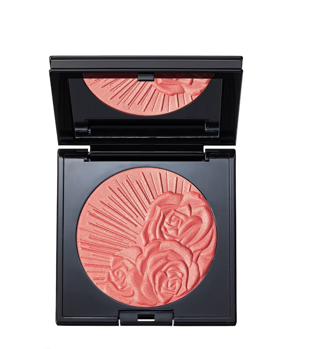 An open black makeup compact showing a round pressed powder blush in a warm coral-pink shade with an embossed rose and sunburst pattern. The compact lid is open, reflecting the blush in the mirror.
